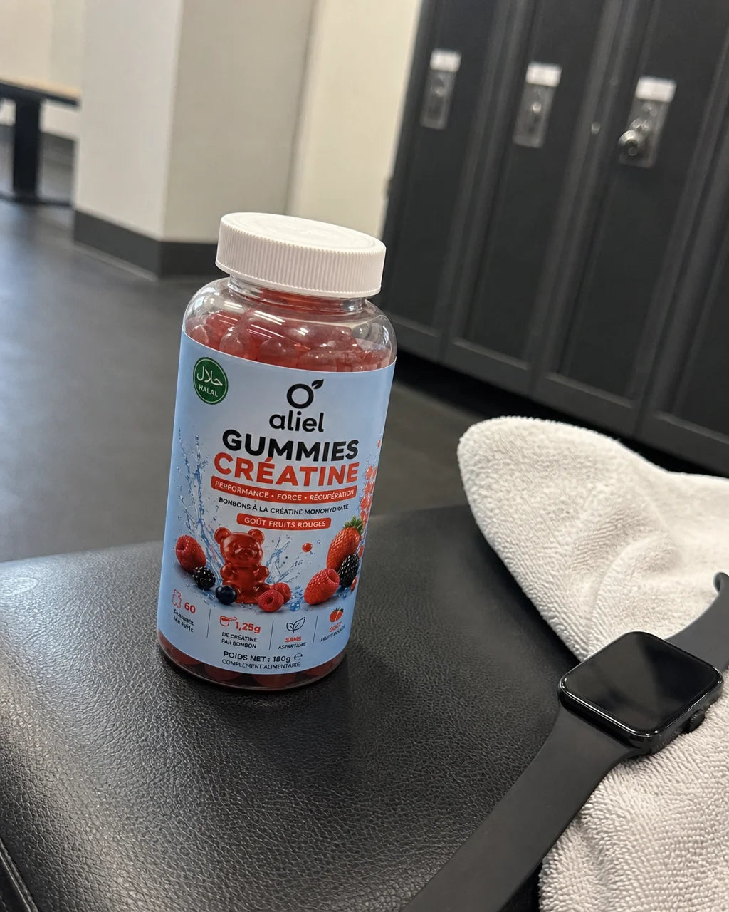 Casual smartphone photograph of the aliel GUMMIES CRÉATINE bottle placed on a gym bench next to a wh